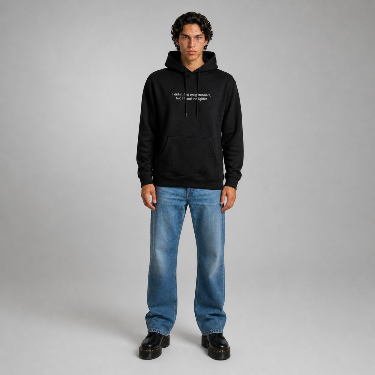 Person wearing a black thick hoodie with text 'I didn't find enlightenment, but I found the lighter' with 400 GSM premium streetwear hoodie for men and women and blue jeans on a gray background.