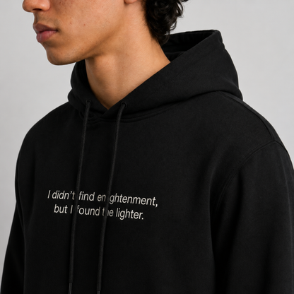 A close up shot of a person wearing a black thick hoodie with text 'I didn't find enlightenment, but I found the lighter' with 400 GSM premium streetwear hoodie for men and women and blue jeans on a gray background.