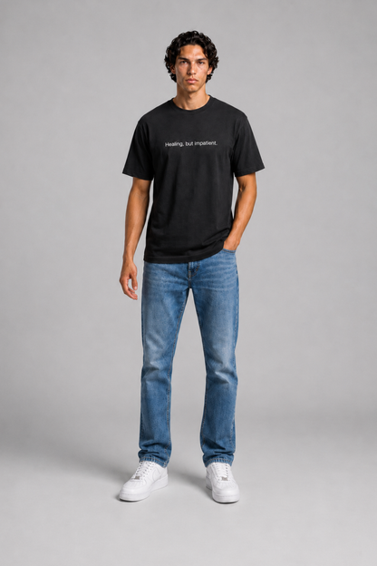 Person wearing a black t-shirt with text 'Healing, but impatient' with 240 GSM premium streetwear t-shirt for men and women and blue jeans on a gray background.