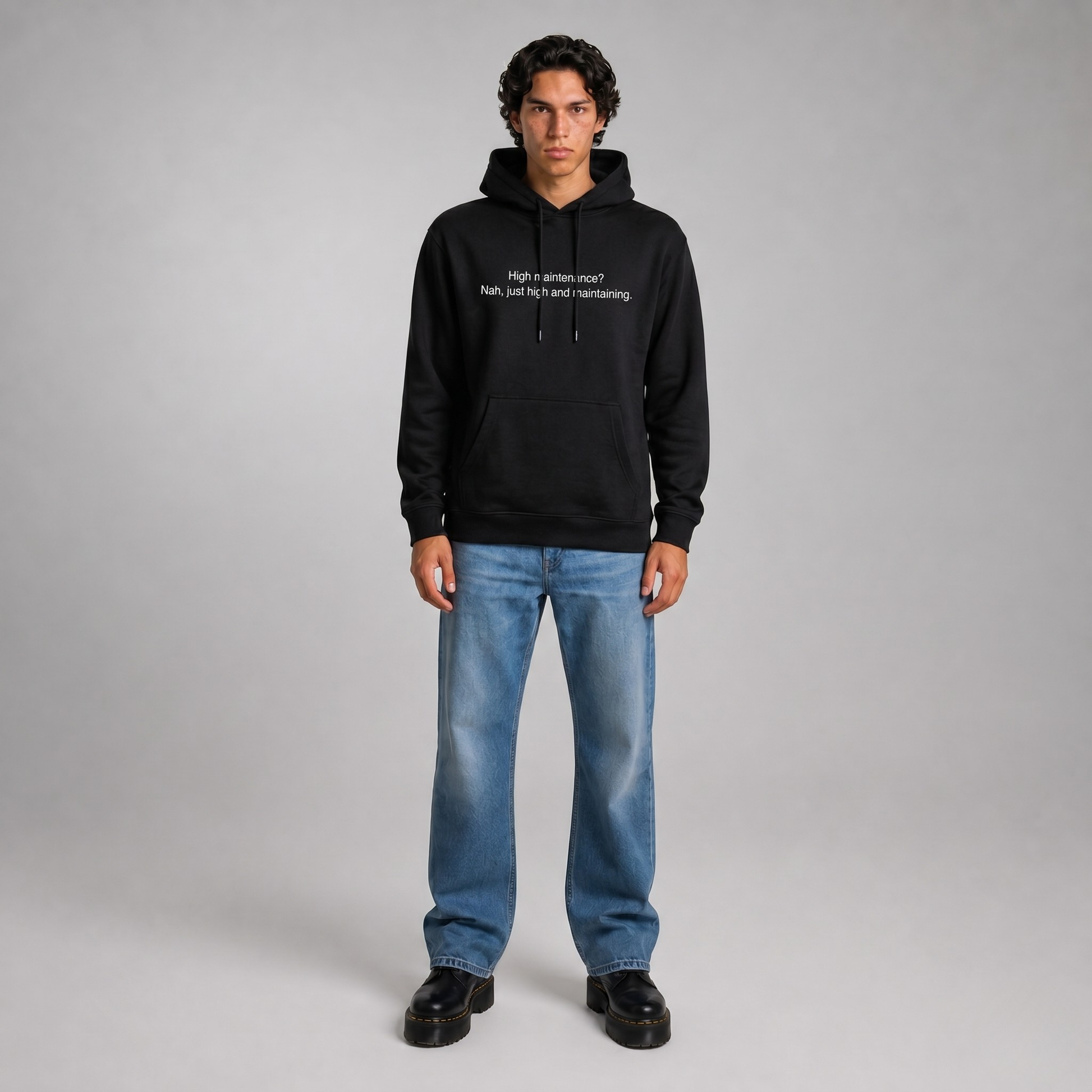 Person wearing a black thick hoodie with text 'High maintenance? Nah, just high and maintaining'  with 400 GSM premium streetwear hoodie for men and women and blue jeans on a gray background