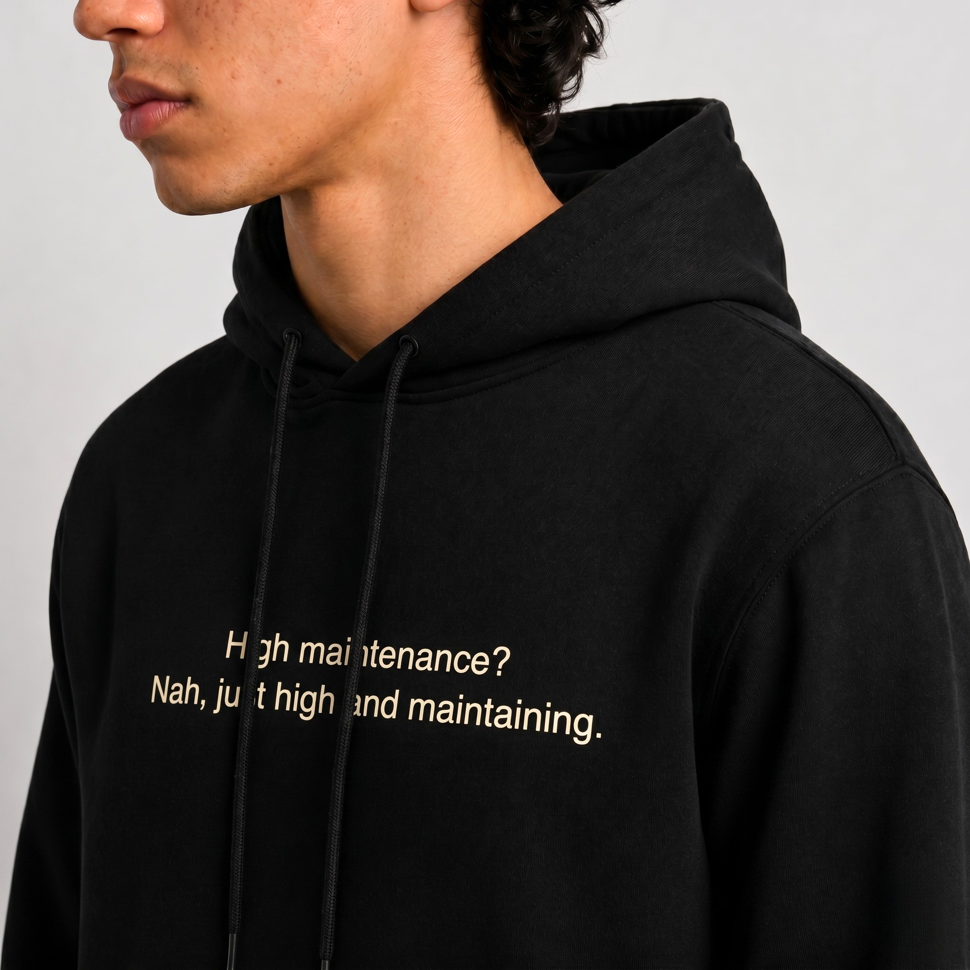 A close up shot of a person wearing a black thick hoodie with text 'High maintenance? Nah, just high and maintaining'  with 400 GSM premium streetwear hoodie for men and women and blue jeans on a gray background.