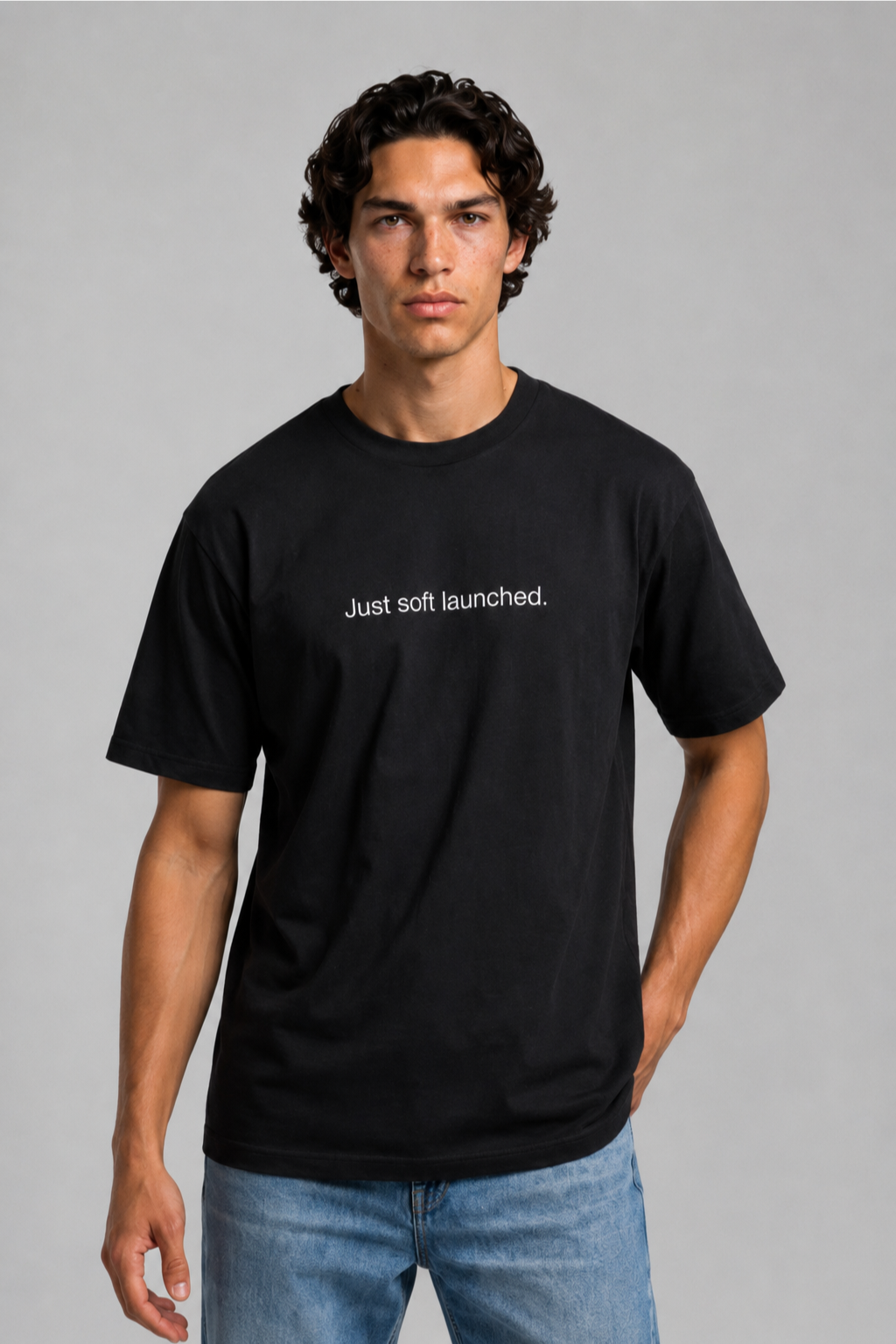A close up shot of a person wearing a black t-shirt with text 'Just soft launched' with 240 GSM premium streetwear t-shirt for men and women and blue jeans on a gray background.