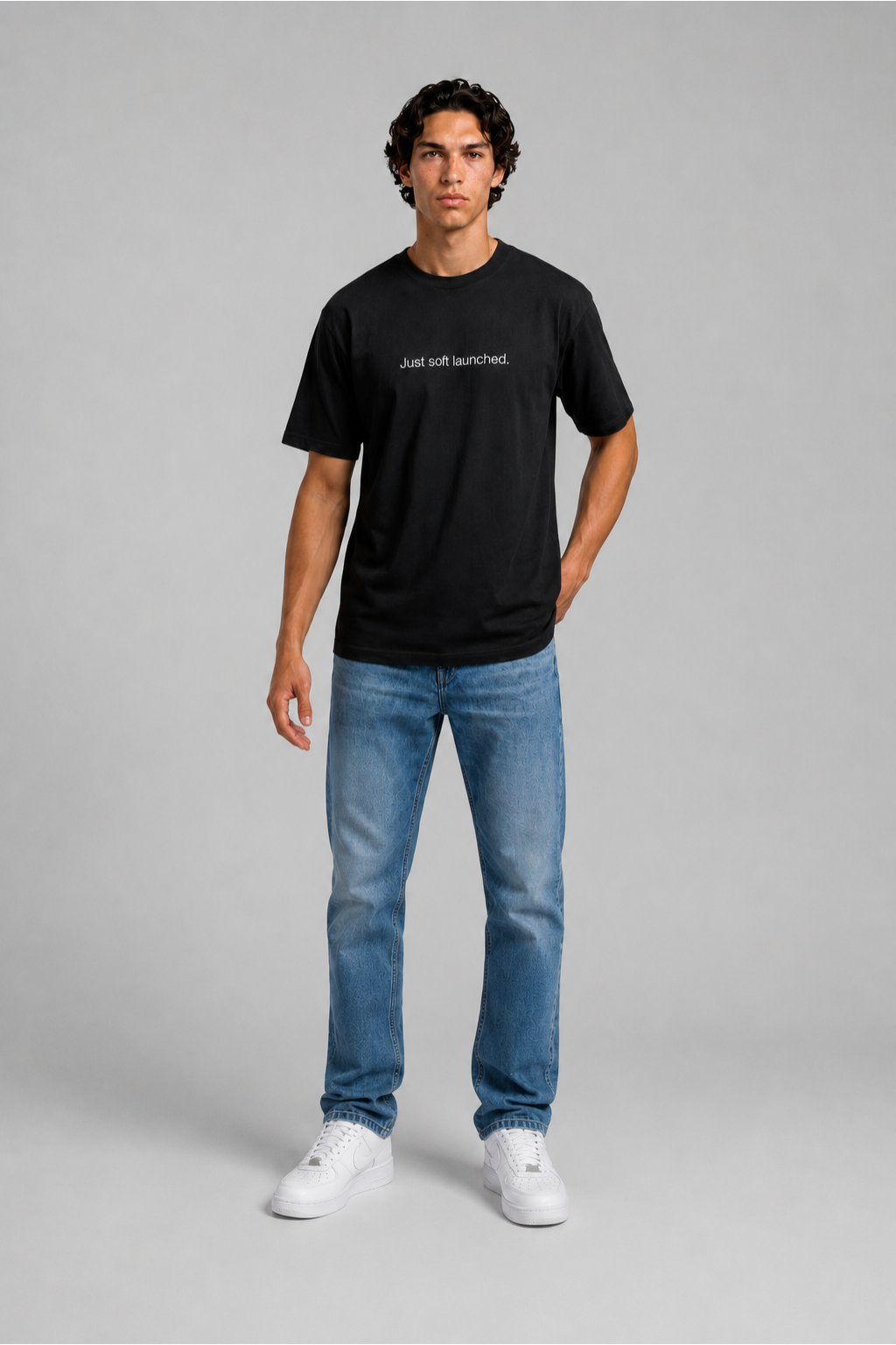 Person wearing a black t-shirt with text 'Just soft launched' with 240 GSM premium streetwear t-shirt for men and women and blue jeans on a gray background.