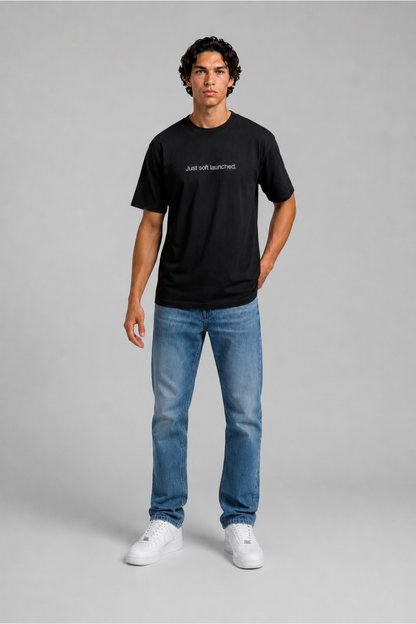 Person wearing a black t-shirt with text 'Just soft launched' with 240 GSM premium streetwear t-shirt for men and women and blue jeans on a gray background.