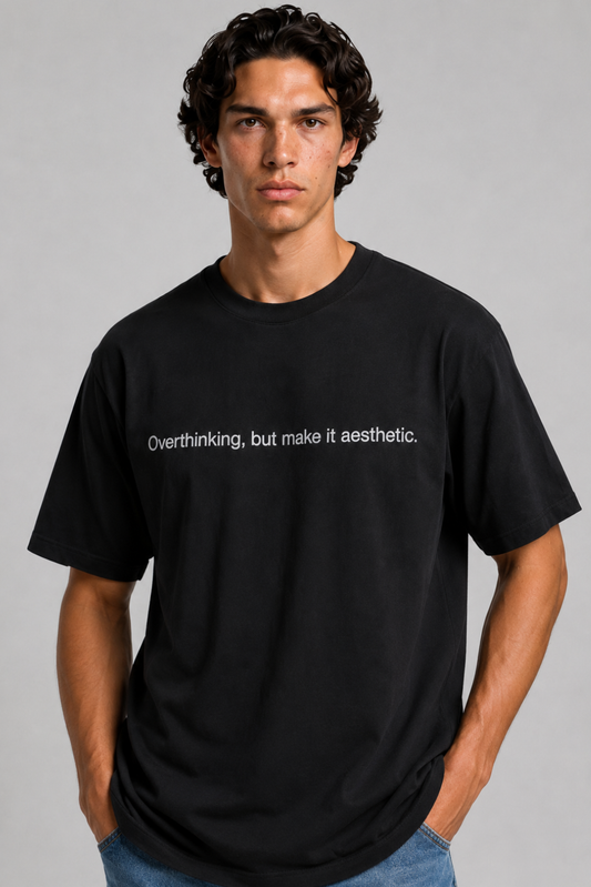 A close up shot of a person wearing a black t-shirt with text 'Overthinking, but make it aesthetic' with 240 GSM premium streetwear t-shirt for men and women and blue jeans on a gray background.