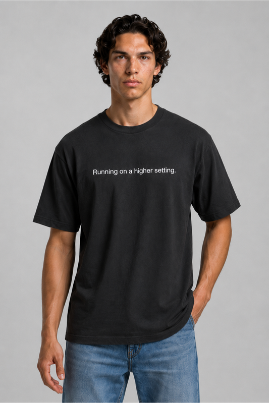 A close up shot of a person wearing a black t-shirt with text 'Running on a higher setting.' with 240 GSM premium streetwear t-shirt for men and women and blue jeans on a gray background.